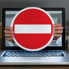 Ręce trzymają znak zakazu wjazdu przed ekranem laptopa, symbolizując cenzurę w internecie.