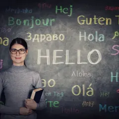 Kobieta z książką na tle tablicy z napisem "HELLO" i powitaniami w różnych językach, symbolizującymi najtrudniejsze języki świata.