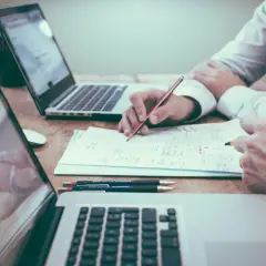 Dwóch mężczyzn pracuje nad dokumentami, używając laptopów. Jeden z nich pisze, tworząc skomplikowany **akronim** na papierze.
