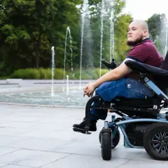 Mężczyzna na wózku inwalidzkim patrzy w dal, siedząc przy fontannie.