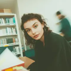 Młoda kobieta studiuje w bibliotece, otoczona regałami pełnymi książek. Jej wzrok skierowany jest na otwartą księgę, a w tle widać inne osoby pochłonięte lekturą. To idealne miejsce do tworzenia bibliografii.