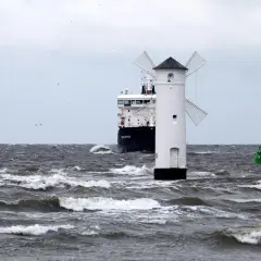 Biała latarnia morska w kształcie wiatraka na wzburzonym morzu, obok statku.