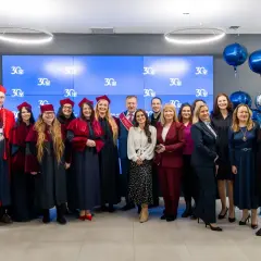 Grupa absolwentów i wykładowców świętuje 30-lecie swojej alma mater. Wszyscy ubrani są w togi i birety, a w tle wiszą balony.