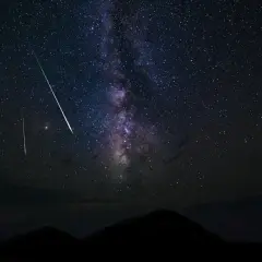 Nocne niebo rozświetla Droga Mleczna i kilka jasnych smug, jakby spadająca gwiazda na tle górskiego krajobrazu.