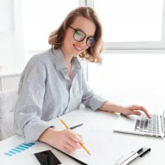 Uśmiechnięta kobieta w okularach pracuje przy laptopie i notuje. Przygotowuje wniosek o dotację.