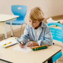 Chłopiec z blond włosami, ubrany w błękitną koszulę, siedzi przy biurku i rysuje. Obok niego leżą kredki, zeszyt i butelka z wodą. Coraz lepiej mu idzie.