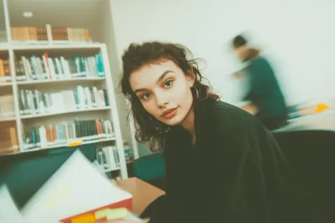 Młoda kobieta studiuje w bibliotece, otoczona regałami pełnymi książek. Jej wzrok skierowany jest na otwartą księgę, a w tle widać inne osoby pochłonięte lekturą. To idealne miejsce do tworzenia bibliografii.