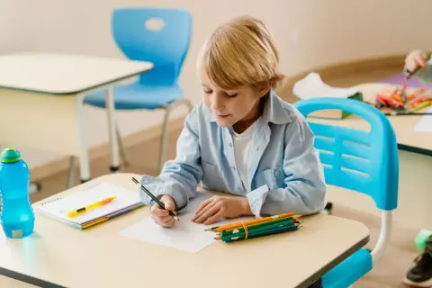 Chłopiec z blond włosami, ubrany w błękitną koszulę, siedzi przy biurku i rysuje. Obok niego leżą kredki, zeszyt i butelka z wodą. Coraz lepiej mu idzie.