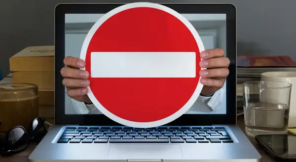 Ręce trzymają znak zakazu wjazdu przed ekranem laptopa, symbolizując cenzurę w internecie.
