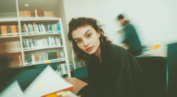 Młoda kobieta studiuje w bibliotece, otoczona regałami pełnymi książek. Jej wzrok skierowany jest na otwartą księgę, a w tle widać inne osoby pochłonięte lekturą. To idealne miejsce do tworzenia bibliografii.