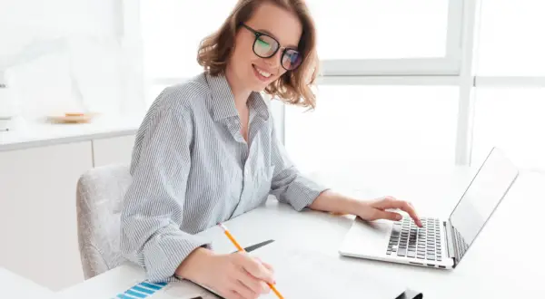 Uśmiechnięta kobieta w okularach pracuje przy laptopie i notuje. Przygotowuje wniosek o dotację.
