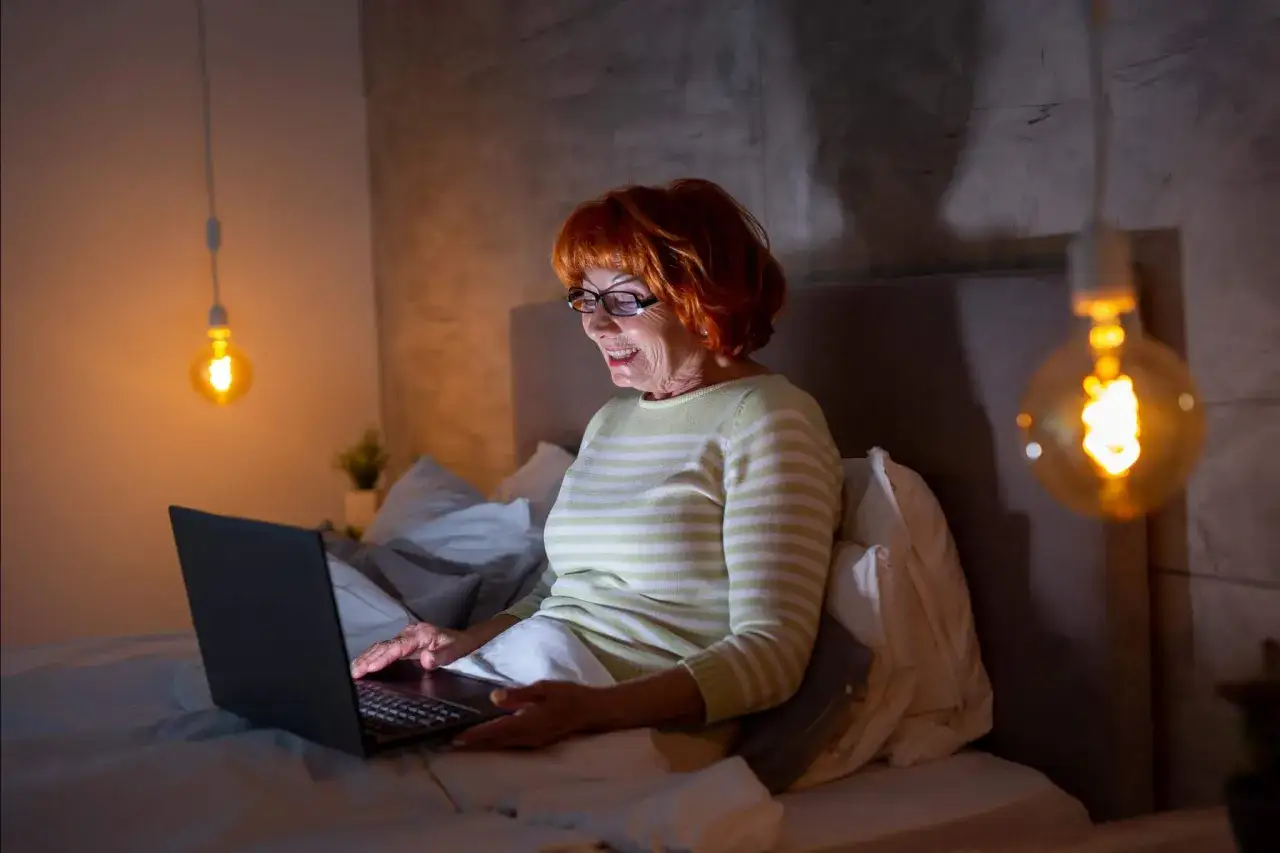 Uśmiechnięta starsza kobieta z rudymi włosami i okularami korzysta z laptopa w łóżku. To dowód, że ageizm jest nieuzasadniony – wiek to tylko liczba.