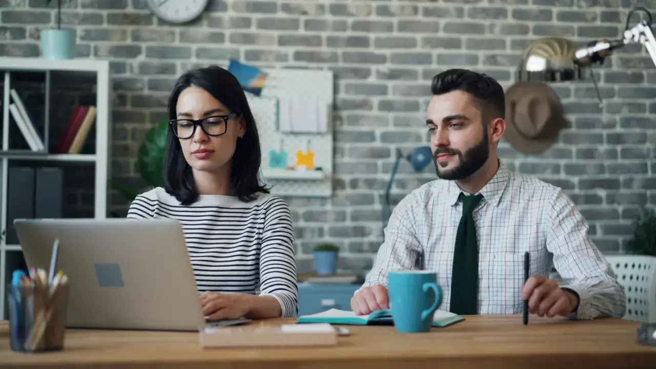Kobieta w okularach pracuje przy laptopie, mężczyzna obok z zaangażowaniem obserwuje ekran.