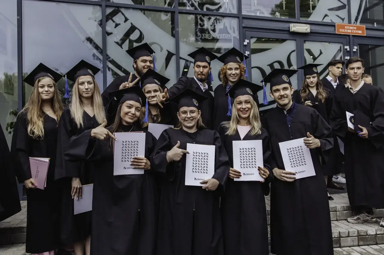 Absolwenci w togach i biretach świętują przed budynkiem swojej alma mater.