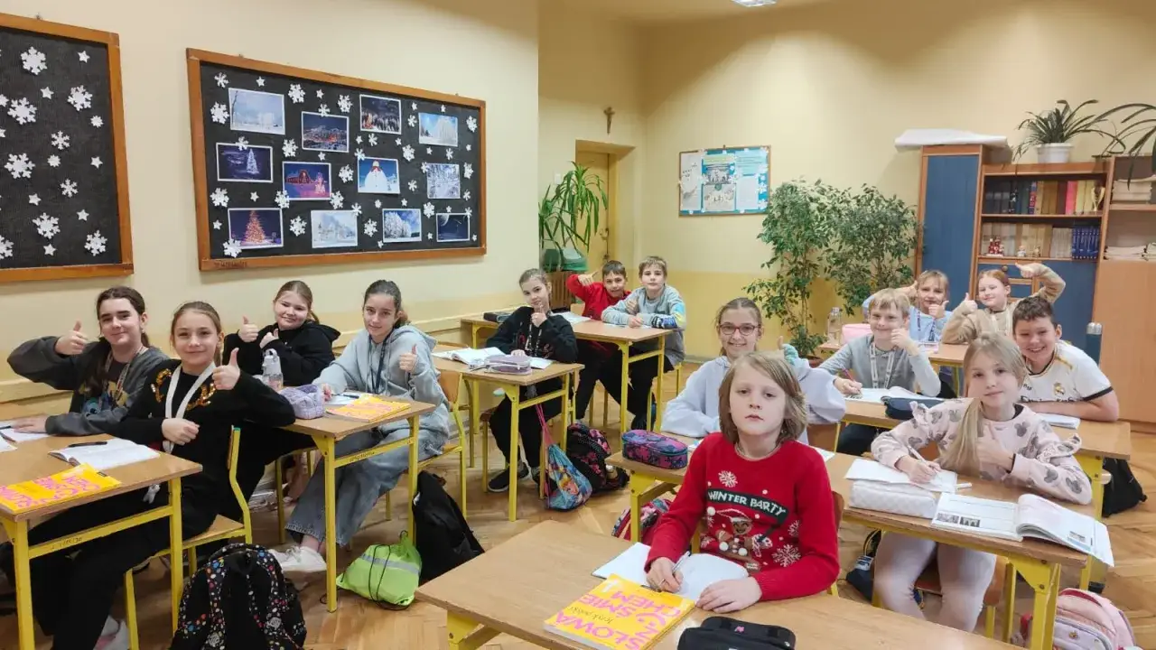 Uczniowie w klasie z uśmiechem wykonują obowiązek szkolny, pokazując kciuki w górę.