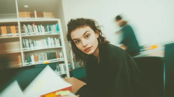 Młoda kobieta studiuje w bibliotece, otoczona regałami pełnymi książek. Jej wzrok skierowany jest na otwartą księgę, a w tle widać inne osoby pochłonięte lekturą. To idealne miejsce do tworzenia bibliografii.